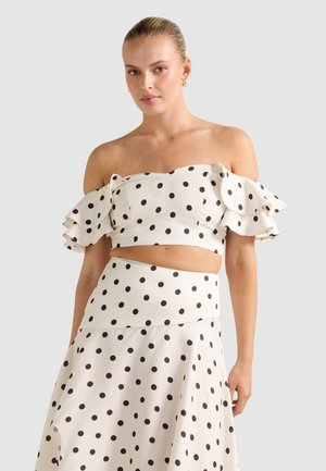 Woman wearing white off-shoulder crop top and matching high-waisted skirt with black polka dots against a plain light background.