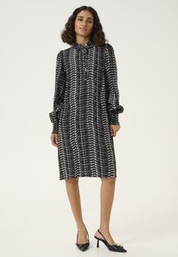 Rochie neagră cu model, cu mâneci lungi și detalii cu nasturi, realizată dintr-un material ușor. Asortată cu pantofi slingback negri.