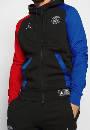 Homme portant un sweat à capuche noir zippé avec une manche gauche rouge, une manche droite bleue, les logos Paris Saint-Germain et Jordan, et un pantalon noir assorti.