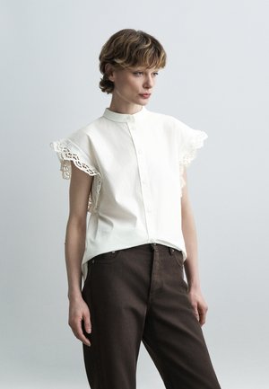 Woman with short hair wearing a white button-up blouse with lace-trimmed sleeves and dark brown pants, standing against a plain light background.