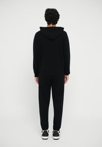 Set cu hanorac negru și pantaloni de trening dintr-un material moale, cu o croială lejeră și manșete cu bandă nervurată. Adidași cu accente contrastante alb-negru.