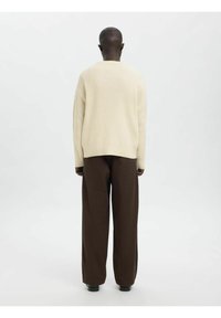 Bež pulover z rebrastim vzorcem in sproščenim krojem, kombiniran z širokimi rjavimi hlačami, prikazan od zadaj na enobarvnem ozadju.