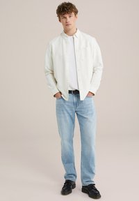 Cămașă albă cu dungi subtile, purtată peste un tricou alb; blugi bleu deschis; pantofi negri; model stând pe un fundal neutru.