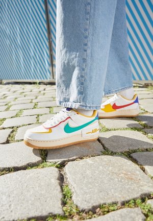 Osoba nosząca białe buty Nike Air z kolorowymi logo i jasnoniebieskie dżinsy stojąca na bruku w pobliżu niebiesko-paskowanego ogrodzenia.