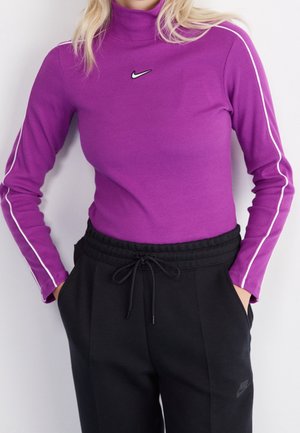 Kobieta w dopasowanym fioletowym golfie z białymi paskami na rękawach, czarne dresy Nike, ręce w kieszeniach, na jednolitym tle.