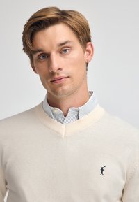 Kremowy sweter z dekoltem w serek wykonany z miękkiego materiału, z granatowym logo na lewej piersi oraz jasnoniebieską i białą koszulą w paski pod spodem.