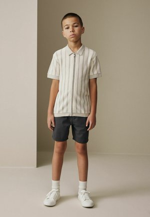 Boy standing against a beige background wearing a short-sleeve zip-up knit shirt, dark shorts, white socks, and white sneakers.