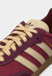 Κοντινό πλάνο αθλητικού παπουτσιού Adidas Samba σε μπορντό χρώμα με μπεζ δερμάτινες ρίγες με ρωγμές, μπεζ κορδόνια, λογότυπο στην γλώσσα και σόλα από καουτσούκ.