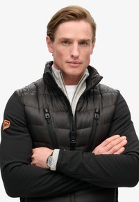 Sort puffy vest med gråt fleecekrave, lynlås-lukninger og orange logo. Modellen har en hvid skjorte og et sort ur på. Armene er korslagte.