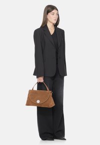 Borsa in suede marrone con chiusura a patta arrotondata e un accento argentato, tenuta da una persona che indossa un blazer nero e pantaloni a gamba larga.