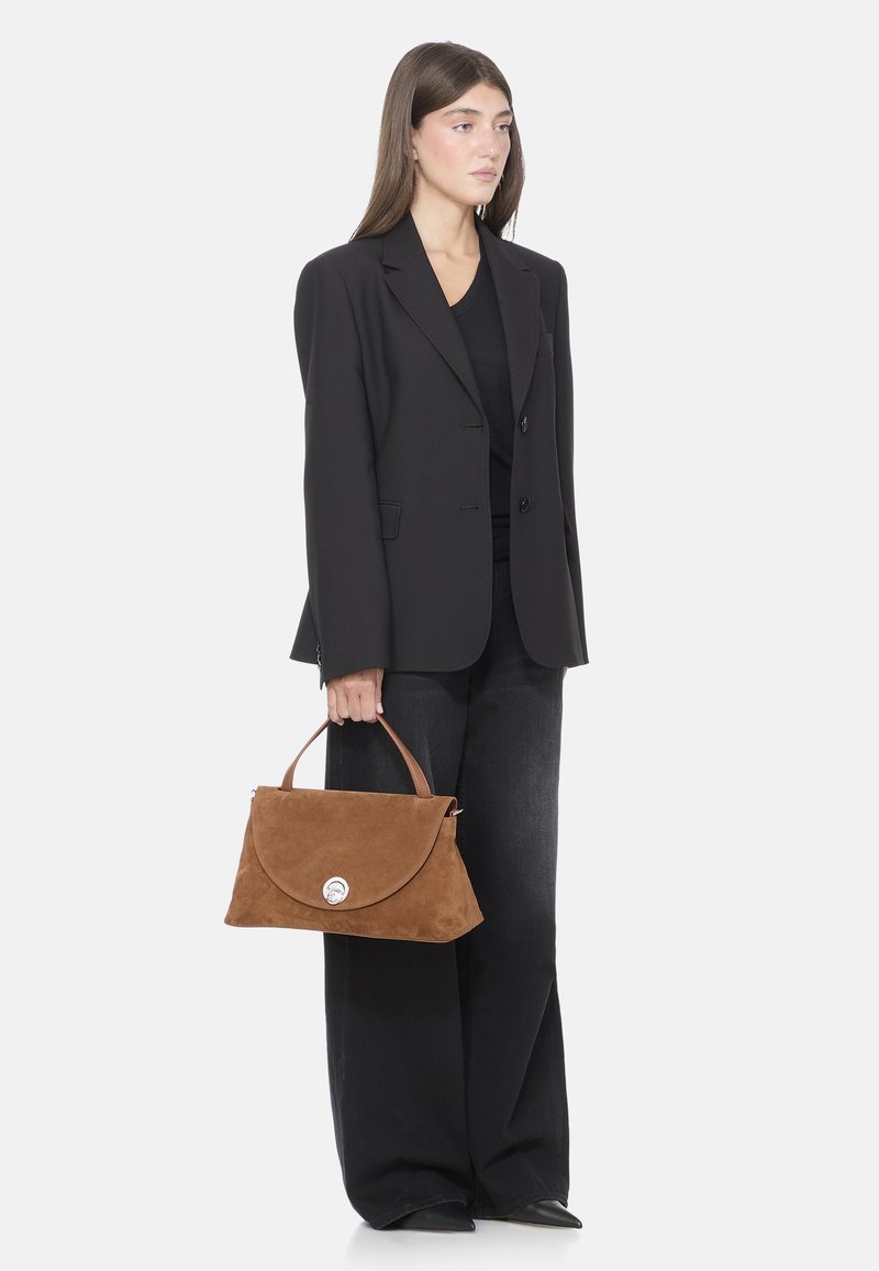 Borsa in suede marrone con chiusura a patta arrotondata e un accento argentato, tenuta da una persona che indossa un blazer nero e pantaloni a gamba larga.