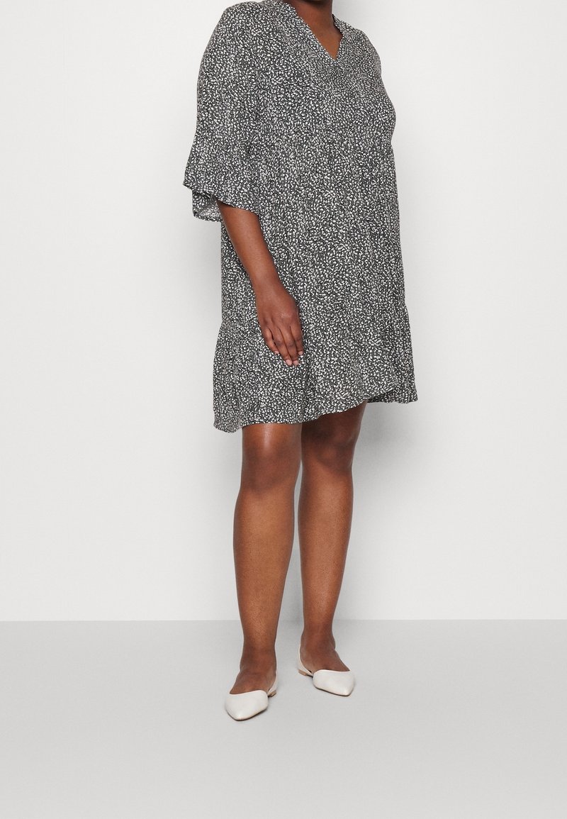 Personne portant une robe ample noire et blanche à motifs, longueur genou, et des chaussures plates blanches pointues, debout devant un fond uni.
