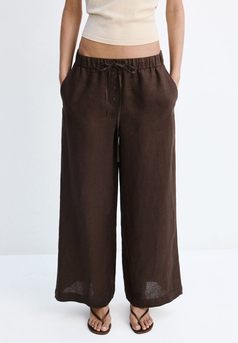 Femme portant un pantalon large marron foncé ample avec une taille à cordon et un détail de boutons, associé à des tongs et un crop top beige.