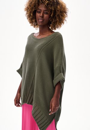 Kobieta o kręconych włosach, ubrana w oversize oliwkowo-zielony sweter z dzianiny z podwiniętymi rękawami i jaskrawo różową spódnicę, stojąca na białym tle.