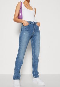 Jeans de tiro alto en un azul claro con adornos plateados dispersos, combinados con una camiseta sin mangas blanca y zapatillas blancas, además de un bolso púrpura.