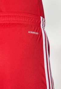 Rode sportieve shorts van zacht materiaal hebben een witte "AEROREADY" logo en drie witte strepen langs de zijkant voor extra detail.