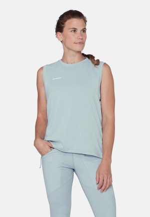 Donna con capelli intrecciati che indossa un top senza maniche azzurro chiaro e pantaloni abbinati, in piedi con una mano in tasca, sfondo bianco.