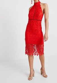 Femme portant une robe halter sans manches en dentelle rouge avec des découpes florales et des sandales à talons beiges, stand contre un fond blanc.