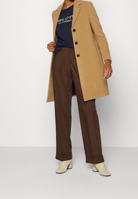 Camelový kabát s přizpůsobeným střihem, černé knoflíky, nošený přes námořnické tričko a hnědé kalhoty s vysokým pasem; doplněný krémovými kotníkovými botami.