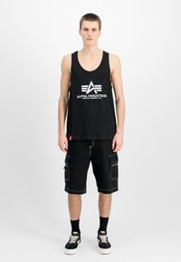Black tank top with white "ALPHA INDUSTRIES" logo, paired with black cargo shorts featuring multiple pockets and white stitching, worn with black sneakers.