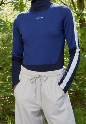 Person wearing a navy blue and black turtleneck top with white stripe on sleeves and light gray drawstring pants, standing outdoors.