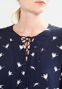 Laivastonsininen paita, jossa on kurkihirvi-printti, jossa on nyöritettävä kaula-aukko ja pehmeä, virtaava kangas. Suunnittelussa on valkoisia ja purppuranvärisiä lintumalleja.