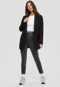 Czarna dopasowana marynarka na białym topie crop, w zestawie z ciemnymi dżinsami. Model stoi z rękami na marynarce, nosząc sneakersy i skarpetki za kostkę.