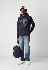 Felpa con cappuccio blu marina e logo bianco, jeans azzurri chiari e sneakers crema. Tiene una borsa scura con estremità tonde e spallacci.