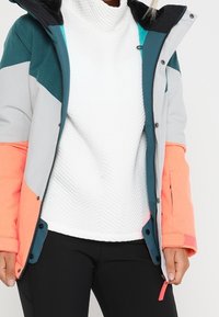 Chaqueta aislante multicolor con paneles en gris, azul verdoso y naranja neón; equipada con un cuello alto y cierre de cremallera.