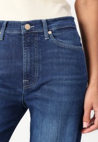 Blå denimjeans med høy midje, med en messingknapp, nagler og sømdetaljer. Glatt tekstur og lett falming ved lårene.