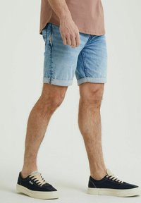 Pantaloni corti in denim azzurro chiaro con orli arrotolati e dettagli strappati, abbinati a sneaker blu scuro e una maglietta beige. Design semplice e casual.