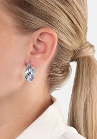 Silver vridet hoopsmycke bärs i vänster öra, ljust blont hår uppsatt, och vit skjorta synlig vid halsringningen.