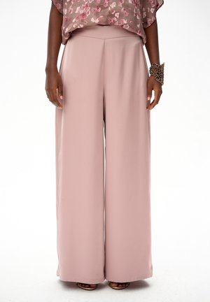 Kobieta ubrana w szerokie spodnie w kolorze różowego brzoskwini, z wysokim stanem, kwiecistą bluzkę z krótkim rękawem oraz duża bransoletkę na lewej ręce.