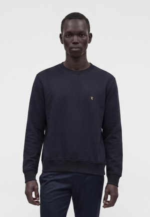 Homme à la peau foncée portant un sweat-shirt uni bleu marine foncé et un pantalon sombre, debout devant un fond gris clair.