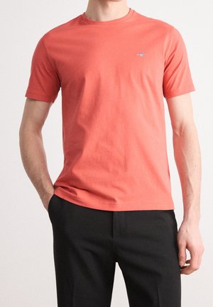 Homme portant un t-shirt corail à manches courtes avec un petit logo sur la poitrine et un pantalon noir, debout avec une main dans la poche.