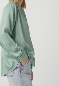 Blusa verde menta con scollatura rotonda, chiusura a bottoni e maniche lunghe. Il tessuto sembra testurizzato, indossata con jeans in denim chiaro.