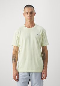 Lys gul bomulds t-shirt med rund hals, korte ærmer og broderet grønt logo på venstre bryst, iført med lyseblå bukser.