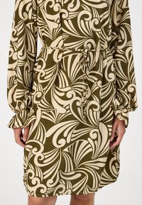 Rochie de culoare verde măsliniu cu un model proeminent de spirale crem, mâneci tip clopot elasticizate și talie cu şnur pentru ajustare. Material neted, ușor.