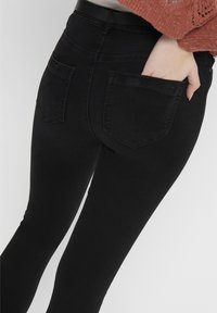 Persona que lleva pantalones ajustados negros con una mano en el bolsillo trasero, combinados con un suéter de punto en color óxido, mostrado de la cintura hacia abajo.