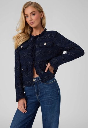Femme blonde portant une veste bleu marine texturée boutonnée avec des poches plaquées et un jean bleu taille haute, posant avec une main sur la hanche.