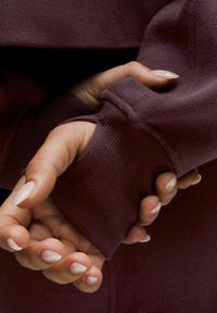 Bordo pulover z rebrastim vzorom na rokavih; roke, ki stisnejo tkanino na zapestju, prikazujejo gladke, negovane nohte.