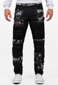 Pantaloni in denim nero con numerosi patch, testo stampato e borchie argentate. Presentano una texture a coste e tasche con zip.