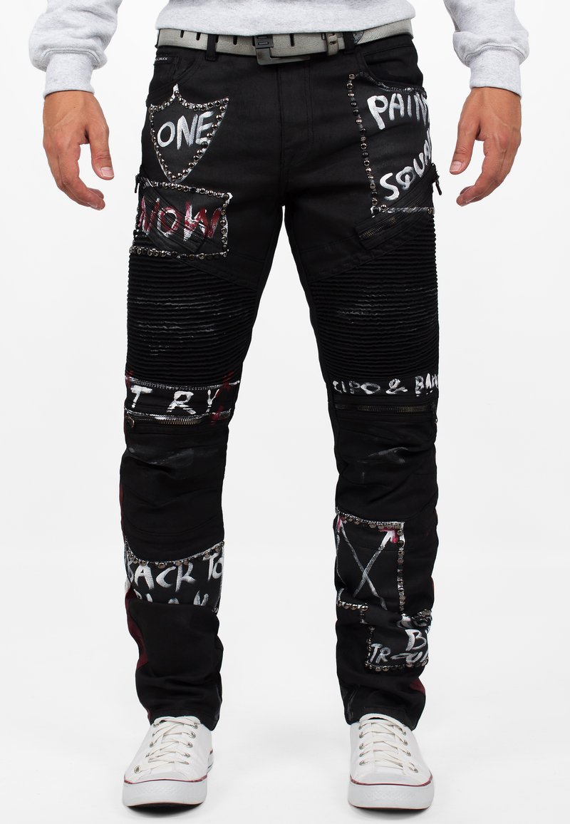 Pantaloni in denim nero con numerosi patch, testo stampato e borchie argentate. Presentano una texture a coste e tasche con zip.