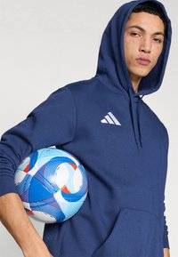 Σκούρο μπλε φούτερ με κουκούλα και τσέπη καγκουρό, με λευκό λογότυπο Adidas, συνοδευόμενο από πολύχρωμη μπάλα ποδοσφαίρου με μπλε, κόκκινα και λευκά σχέδια.