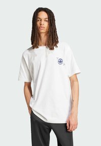 Bijela pamučna majica s okruglim izrezom, kratkim rukavima i plavim logotipom na lijevoj strani prsnog koša. Uparena s tamnim hlačama, stoji naslonjena na svijetlu pozadinu.