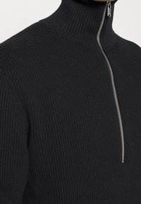 Maglione nero a coste con collo alto, caratterizzato da una cerniera argentata sul davanti, con una finitura in tessuto texturizzato e una vestibilità aderente attorno al collo.