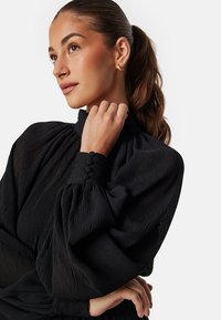 Blouse noire texturée avec un col haut froncé et de longues manches bouffantes. Présente des embellissements en boutons près des poignets.