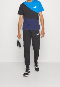 Tričko s barevnými bloky Puma v modré, námořnické a černé barvě, spárované s černými kalhotami. Osoba drží švihadlo a má na sobě černé tenisky.