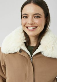 Veste marron à fermeture éclair avec un col en fausse fourrure blanc, associée à un pull vert. La texture semble douce et chaude.