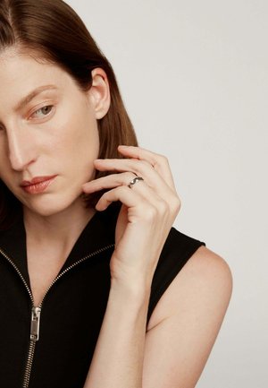 Mujer con cabello corto y castaño, vestida con una blusa sin mangas negra con cremallera, tocándose la cara y mostrando un anillo plateado en la mano derecha.